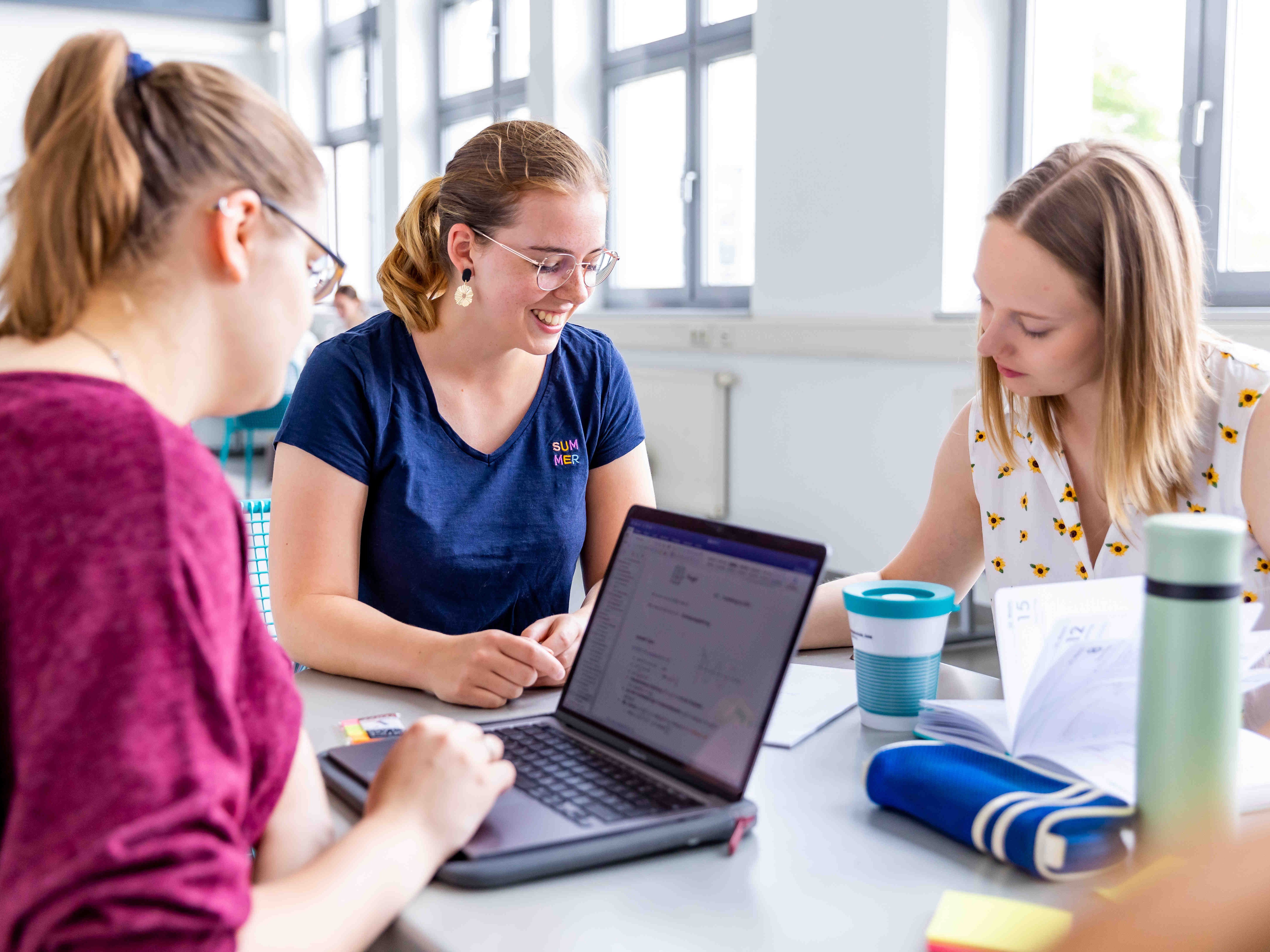 Drei junge Frauen sitzen an einem Tisch, eine davon arbeitet am Laptop.