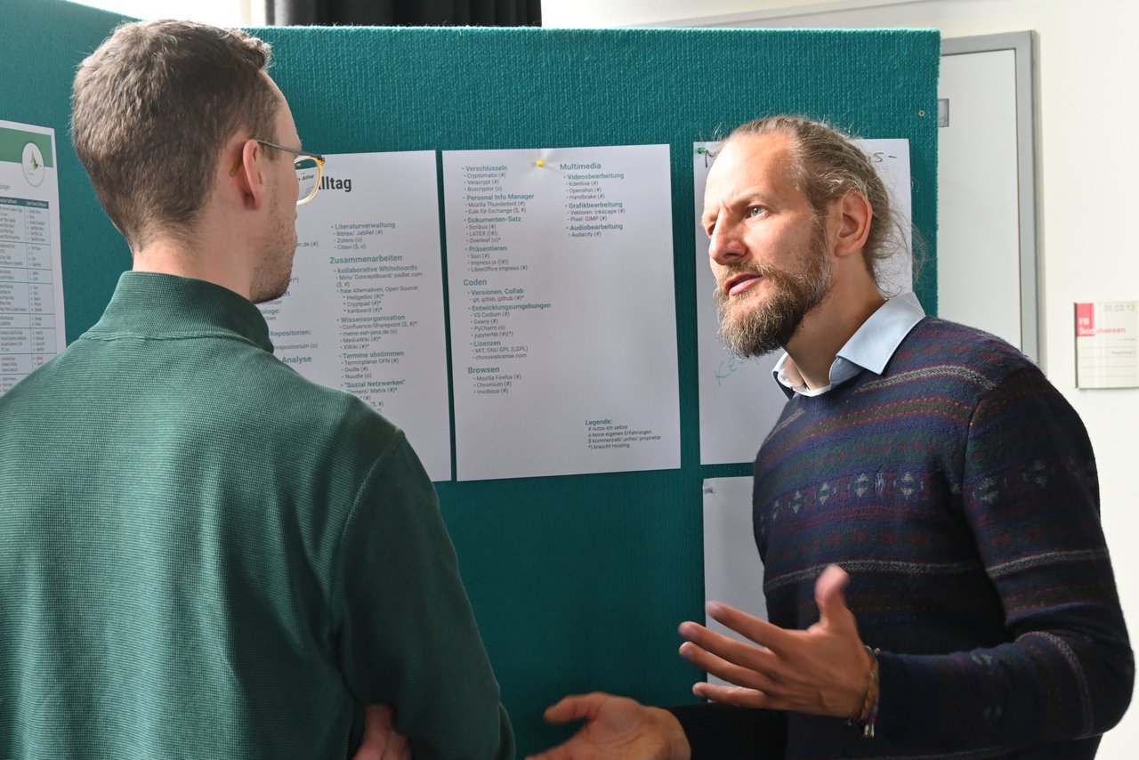 Zwei Männer beim intensiven Austausch vor einer Posterwand, am Thementisch zu Software für den Datenalltag